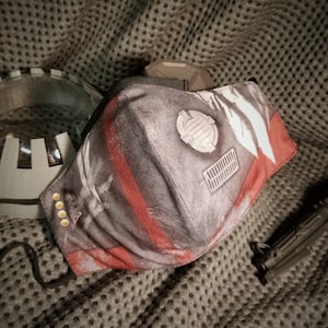 May include: A gray and red face mask with a weathered, industrial design. The mask features a series of red stripes, a circular emblem, and a rectangular vent design. Gold-colored rivets are visible on the side. A black cord is attached for securing the mask.