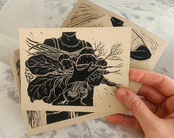 May include: Several square cards with a linocut print of a person holding root vegetables. The print is in black and white and features beets, carrots, and leafy greens. The cards are tan-coloured and held in a hand, with a patterned fabric in the background.