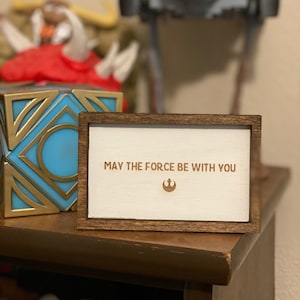 Puede incluir: Un pequeño letrero enmarcado en madera con el texto "MAY THE FORCE BE WITH YOU" y un símbolo de Star Wars. El letrero tiene un fondo blanco y un marco marrón. Otros artículos temáticos de Star Wars están en el fondo.