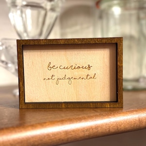 Op de afbeelding: Een klein houten bord met de woorden "be curious not judgmental" gegraveerd op een lichtgekleurd houten paneel. Het frame is donkerbruin van kleur. Het bord staat op een houten plank.