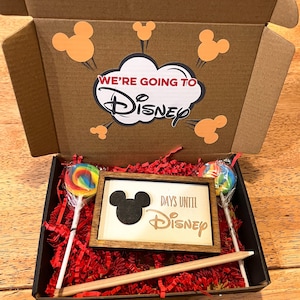 May include: A brown cardboard box with a red and white "We're going to Disney" sign on the top. Inside the box is red tissue paper, a wooden sign with a black Mickey Mouse silhouette and the text "Days until Disney", a rainbow lollipop, and a wooden stick.