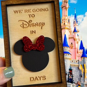 Puede incluir: Un letrero de madera con marco marrón y el texto "We're going to Disney in DAYS" con una silueta negra de la cabeza de Mickey Mouse con un lazo rojo de brillantina.
