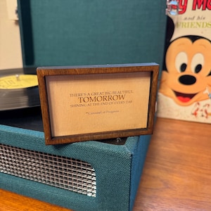 Puede incluir: Un letrero enmarcado de madera con fondo marrón y texto blanco. El texto dice "There's a great big beautiful tomorrow shining at the end of every day." La cita se atribuye a "Carousel of Progress".