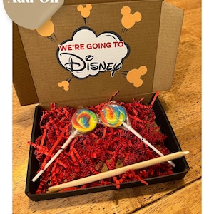 以下が含まれることがあります： 「We're Going to Disney」のステッカーが貼られた段ボール箱の中に、カラフルなロリポップが2本入っています。箱は赤いシュレッダーペーパーでいっぱいです。ロリポップは白い棒についています。追加はディズニーカウントダウンサインと一緒に購入する必要があります。