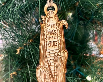Puede incluir: Un adorno de maíz de madera con el texto "It has the juice" colgado de un árbol de Navidad.