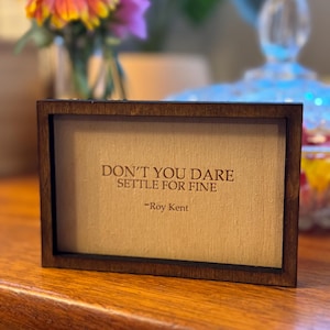 May include: A wooden framed sign with the quote "Don't you dare settle for fine - Roy Kent" in a brown font.