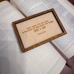 Puede incluir: Una placa enmarcada en madera con la cita "They think I do not know a buttload of crap about the Gospel, BUT I DO!" de Nacho Libre. La placa descansa sobre una Biblia abierta, con el texto de Lucas visible.