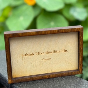 Puede incluir: Un letrero de madera con marco de madera oscura y fondo beige. El texto "I think I like this little life. - Cordelia" está grabado en marrón.