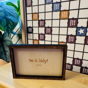 Puede incluir: Un letrero enmarcado en madera con el texto "Do it lady!" y el nombre "-Chil" en una fuente marrón claro. Un tablero de Scrabble está en el fondo. El marco es de color marrón oscuro.