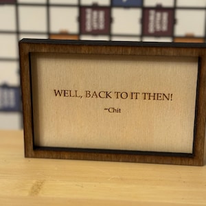 May include: A small, rectangular wooden frame with the text "WELL, BACK TO IT THEN!" and the name "-Chit" printed on a beige background. The frame is dark brown and sits on a light brown surface.