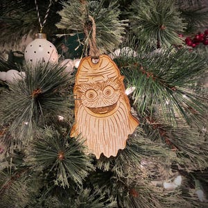 Könnte beinhalten: Ein helles Holzornament mit einer lasergravierten Illustration eines pelzigen Wesens mit großen runden Augen, einem breiten Mund und einem strukturierten Bart. Das Ornament ist an einer dünnen Schnurschlaufe aufgehängt und flach.
