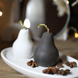 Op de afbeelding: Twee peer-vormige betonnen kaarshouders, een wit en een zwart, met gouden draadstengels. De kaarshouders staan op een wit ovaal dienblad met steranijs peulen.