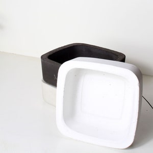 Peut inclure: Deux pots carrés en béton, l'un noir et l'autre blanc. Le pot blanc est posé sur le pot noir.