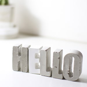 Könnte beinhalten: Ein Satz Betonbuchstaben, die das Wort "HELLO" in Weiß und Grau buchstabieren. Die Buchstaben sind in einer Reihe auf einer weißen Oberfläche angeordnet.