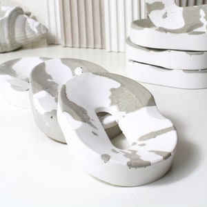 Op de afbeelding: Drie witte en grijze ringvormige objecten van beton met een gat in het midden. De objecten zijn op elkaar gestapeld. De objecten zijn gemaakt van beton en hebben een ruwe textuur.