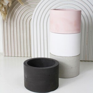 Op de afbeelding: Een verzameling cilindrische plantenbakken in verschillende kleuren. Eén is zwart, een andere is een stapel wit, grijs en roze. De plantenbakken zijn gemaakt van beton en geschikt voor binnen of buiten.