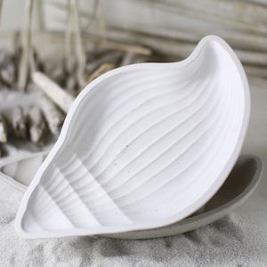May include: White, leaf-shaped decorative dish with a textured, ridged surface. The dish is resting on top of a similar, gray dish. The background features sand and driftwood, suggesting a coastal theme.