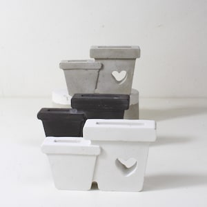 Può includere: Una collezione di piccole fioriere quadrate in cemento nei toni del grigio, nero e bianco. Ogni fioriera ha un'apertura rettangolare nella parte superiore e un ritaglio a forma di cuore sul lato. Le fioriere sono impilate.