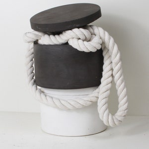 Könnte beinhalten: Ein weißer zylindrischer Behälter mit einem schwarzen Deckel und einem weißen Seil, das darum gewickelt ist. Das Seil ist oben auf dem Behälter zu einem Knoten gebunden.