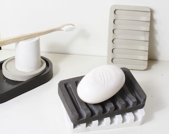 Könnte beinhalten: Eine weiße Seife liegt auf einem dunkelgrauen Seifenschälchen mit weißem Sockel. Das Seifenschälchen ist aus Beton und hat ein geriffeltes Design. Im Hintergrund sind eine Zahnbürste und ein weißer Zahnputzbecher aus Beton zu sehen.