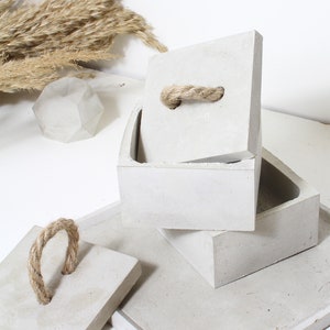 Peut inclure: Trois boîtes en béton avec des poignées en corde. Les boîtes sont empilées les unes sur les autres. La boîte du haut est ouverte et la poignée en corde est visible. Les boîtes sont de couleur gris clair.