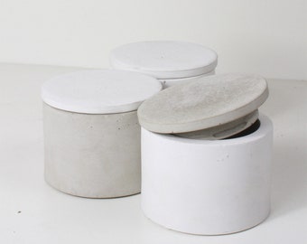May include: Three round concrete containers with lids. Two containers are gray and one is white. The lids are all white.