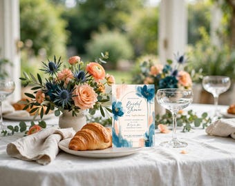 Op de afbeelding: Een rechthoekige uitnodigingskaart voor een bruidsdouche met blauwe en perzikkleurige aquarel bloemenillustraties, gecentreerde tekst en een boogframe, weergegeven op een tafel met een croissant, bloemen en glaswerk.