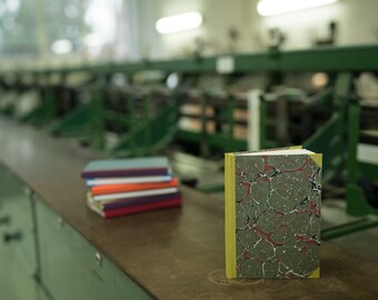 Op de afbeelding: Een stapel kleurrijke notitieboekjes en een enkel, gesloten boek met een marmeren omslag en een gele rug op een groen oppervlak.