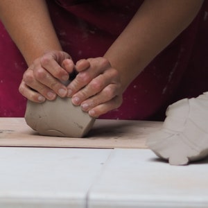 Könnte beinhalten: Die Hände einer Person formen einen Klumpen grauen Tons zu einem Topf auf einer Holzoberfläche. Auf der Oberfläche rechts liegt ein weiteres Stück Ton.