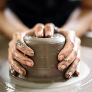 Könnte beinhalten: Die Hände einer Person formen einen Klumpen Ton auf einer Töpferscheibe. Der Ton ist hellbraun und wird zu einer runden Form geformt.