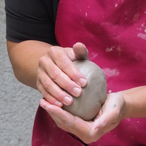 Könnte beinhalten: Die Hände einer Person formen eine Kugel aus grauem Ton. Der Ton ist glatt und hat eine leicht strukturierte Oberfläche. Die Person trägt eine rosa Schürze.