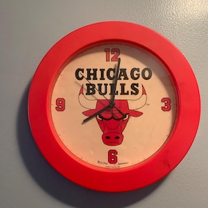 May include: Red round wall clock with a white face featuring the Chicago Bulls logo and the words "Chicago Bulls" in black. The clock has black hands and black numbers.