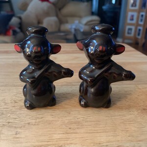 May include: A pair of brown ceramic salt and pepper shakers shaped like mice playing musical instruments. The mice have black eyes, pink noses, and red cheeks. They are wearing black hats and holding brown instruments.