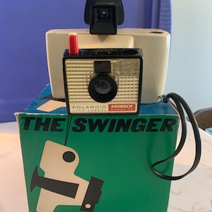 May include: Vintage Polaroid Swinger Land Camera Model 20, with a black lens, red button, and a black viewfinder. The camera is on top of its original green box, which reads "THE SWINGER" in white text.