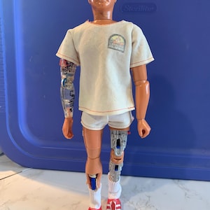 May include: A vintage action figure of a man in a white t-shirt and shorts, with a patch that reads "Six Million Dollar Man". The figure has a bionic arm and leg, red shoes, and white socks. The figure is approximately 12 inches tall.