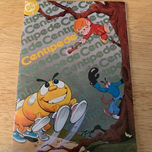 May include: A vintage comic book titled "Centipede" featuring cartoon illustrations. The cover displays a yellow centipede, a red-clad character, and a blue character. The DC Comics logo is visible in the upper left corner. The background has the word "Centipede" repeated.