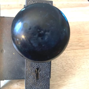 May include: Antique black door knob with a round, glossy finish. The knob is mounted on a decorative, rectangular metal plate with an ornate, swirling pattern and a keyhole. The hardware is likely vintage.