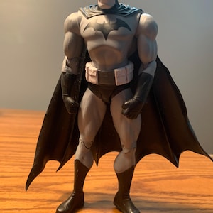 May include: A Batman action figure in a gray and black color scheme. The figure has a bat symbol on the chest, a black cape, and boots. The figure is standing on a wooden surface.