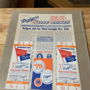May include: Two vintage Los Angeles Dodgers baseball tickets from 1979. The tickets are for the opening day game against the San Diego Padres. The tickets are blue, orange, and white with the Dodgers logo and the text "Opening Day" and "Two Consecutive National League Championships".