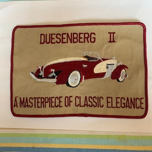 May include: A beige embroidered patch with a red border featuring a red and white Duesenberg II car with the text "DUESENBERG II A MASTERPIECE OF CLASSIC ELEGANCE".