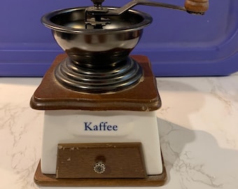 May include: A manual coffee grinder with a white ceramic base featuring the word Kaffee in blue text. It has a dark brown wooden top and a matching wooden drawer with a small knob. The top assembly is black metal with a wooden handle.