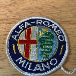 May include: Embroidered patch featuring the Alfa Romeo logo. The circular patch has a navy blue border with the words "ALFA-ROMEO MILANO" in white. The logo inside includes a red cross and a green serpent.