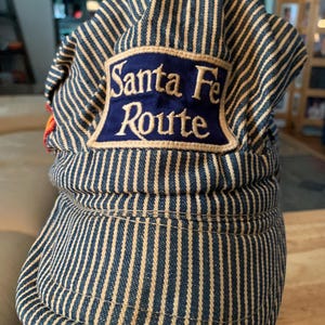 May include: A blue and beige striped railroad cap with the words "Santa Fe Route" embroidered on a navy blue patch. The cap has a rounded crown and a short brim, and is made of a durable fabric.