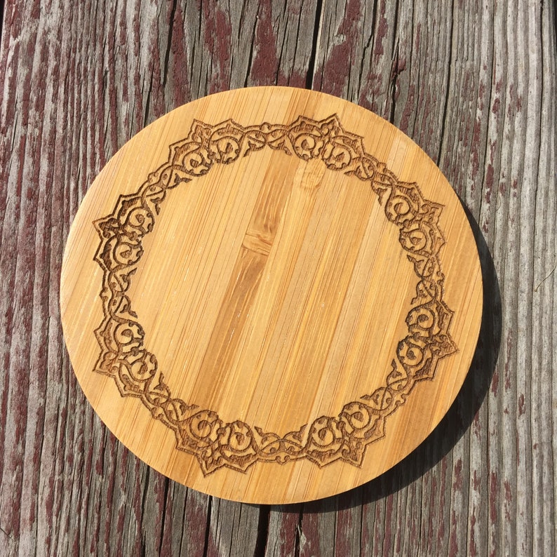 May include: A round wooden coaster with a decorative, intricate design carved into the surface. The coaster is made of light brown wood and has a natural finish.