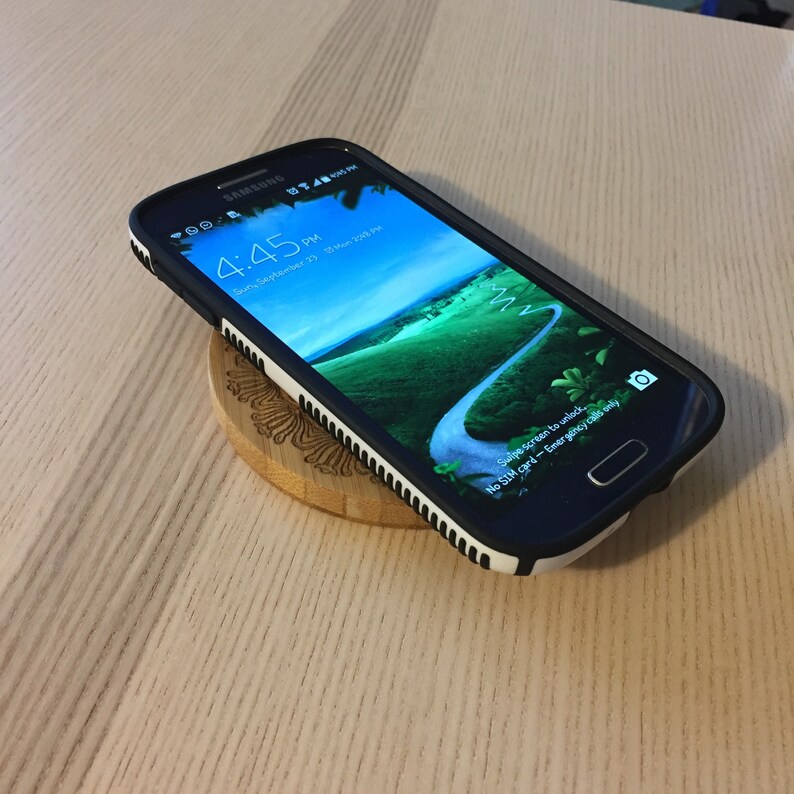 May include: A black Samsung smartphone with a white and black case is charging on a wooden wireless charging pad. The phone's screen displays the time, date, and a message indicating that there is no SIM card inserted.