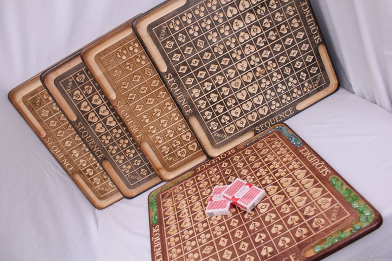 May include: Five wooden game boards with the word "SEQUENCE" printed on them. The boards are made of different types of wood and have a variety of colors. The boards are all the same size and shape. There are playing cards on one of the boards.