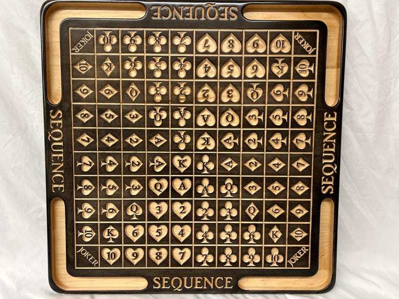 May include: A wooden game board with a black playing surface and a light wood frame. The board is marked with the numbers 2 through 10, the letters J, Q, K, and A, and the word "JOKER". The board is designed for the game "SEQUENCE".