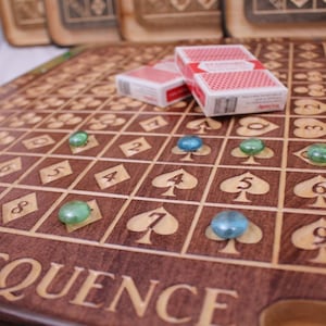 Peut inclure: Un plateau de jeu en bois avec le mot "SEQUENCE" gravé en bas. Le plateau a une grille avec des numéros et des symboles de cartes à jouer. Il y a des pions de jeu en verre vert et bleu sur le plateau. Deux paquets de cartes à jouer sont sur le plateau.