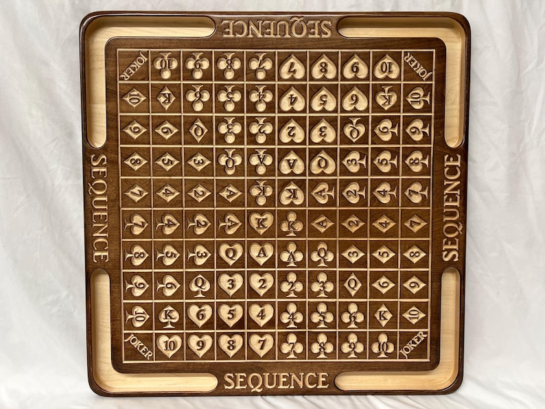 May include: A wooden game board with a grid of 100 squares, each containing a playing card symbol. The board is labeled "SEQUENCE" on all four sides. The board is made of dark brown wood with a light brown border.