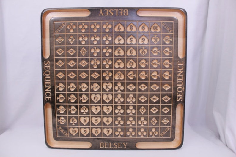 Pu&ograve; includere: Un tabellone da gioco in legno con una superficie marrone scuro e un bordo marrone chiaro. Il tabellone &egrave; contrassegnato da una griglia di quadrati, ognuno dei quali contiene un simbolo di carta da gioco. Il tabellone &egrave; etichettato "SEQUENCE" sui lati sinistro e destro e "BELSEY" in alto e in basso.